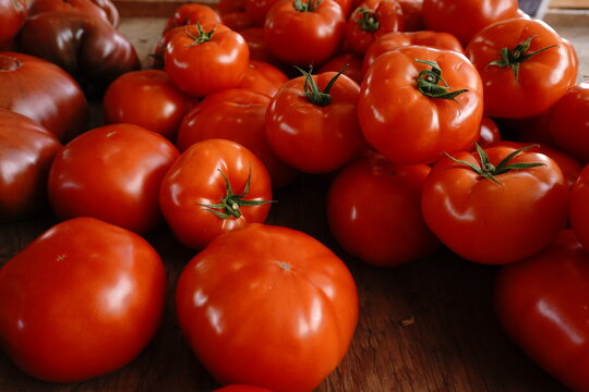 A close-up eye-level view of ripe red tomatoes piled on a rustic wooden surface. Their smooth glossy skins reflect the light highlighting their fresh natural appearance