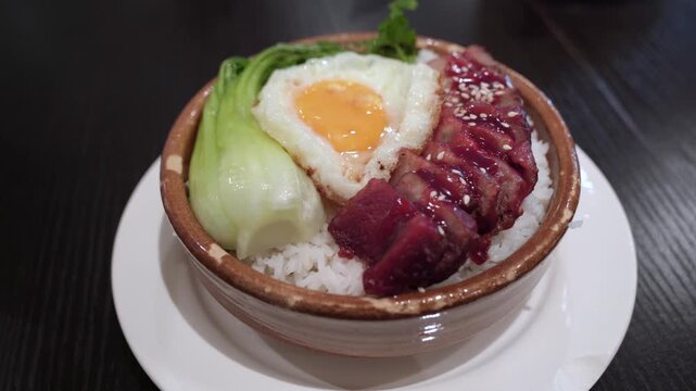 Close-up of a traditional Chinese BBQ pork rice bowl served in a ceramic bowl, featuring sliced char siu pork, a sunny-side-up fried egg, steamed bok choy, and white rice, topped with sesame seeds.