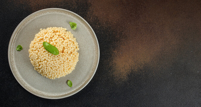 Plate of cooked couscous garnished with fresh basil leaves on a textured dark background, showcasing the fine grains and culinary presentation. top view. copy space.