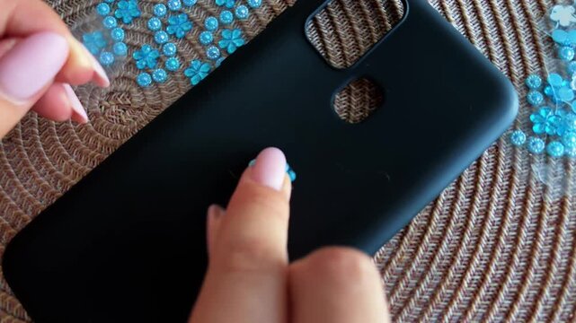 Woman carefully peeling off a blue rhinestone flower sticker from a matte black smartphone case, revealing existing scratches and wear underneath the decoration