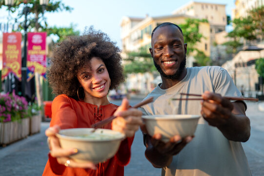 Young couple embracing the trend of Asian cuisine, happily eating noodles with chopsticks outdoors