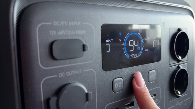 Woman's finger turning off a portable power station with digital display, showing dc and ac output, usb ports, and solar panel input for off grid energy storage