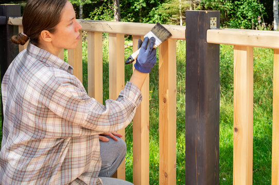 Close up side view of a focused woman applying protective oil coating to a wooden terrace railing with a paintbrush. DIY home improvement and deck maintenance.