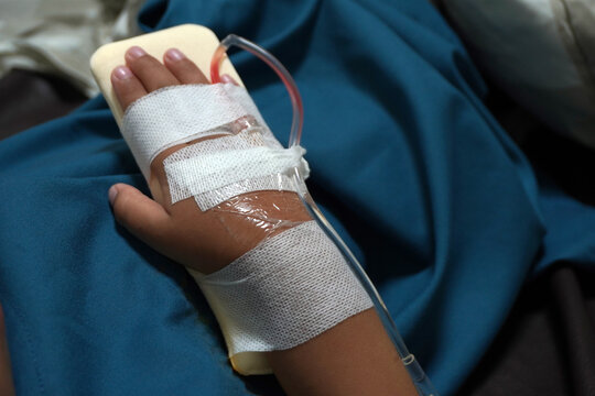 Close-up photo of a patient's hand resting on a padded splint in a hospital bed. The hand has an intravenous (IV) catheter inserted and secured with medical bandages and tape. 