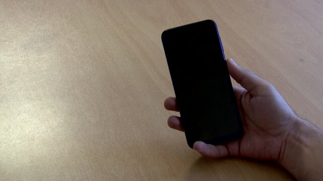 High-resolution 4K close-up video of a hand picking up a sleek black Android smartphone from a wooden table, unlocking it, scrolling over a chroma key green screen, locking the device, and placing it 