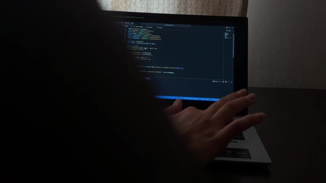 Unrecognizable hacker typing programming code on a laptop keyboard in a dark room. Information technology specialist working on software development and website coding