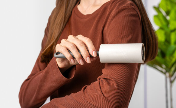 Close up woman hand using sticky adhesive lint roller with paper sheets removing lint from brown clothing at home. Concept of clothing maintenance tool and everyday textile care cleaning routine