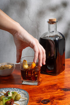 Hand holding a glass of vermouth on the rocks above a wooden table surrounded where there is a bottle, two gildas and a small bowl of peanuts