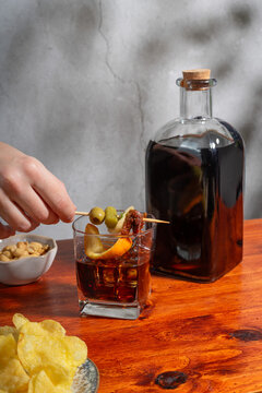 Hand holding gilda on a glass of vermouth on the rocks above a wooden table with a small bowl of peanuts and a plate with crisps
