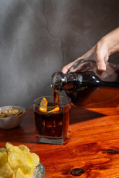 Hand serving a glass of vermouth on the rocks above a wooden table with a small bowl of peanuts and a plate with crisps
