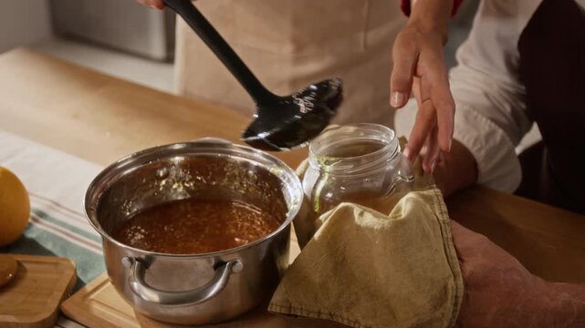 Caucasian woman ladling homemade orange jam into glass jar held by senior Caucasian man at kitchen table. Ideal for culinary tutorials, family lifestyle campaigns, and canning guides