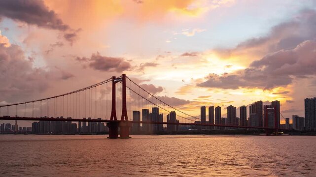 A beautiful suspension bridge spans the river in Wuhan China during a vibrant sunset with a city skyline in the background