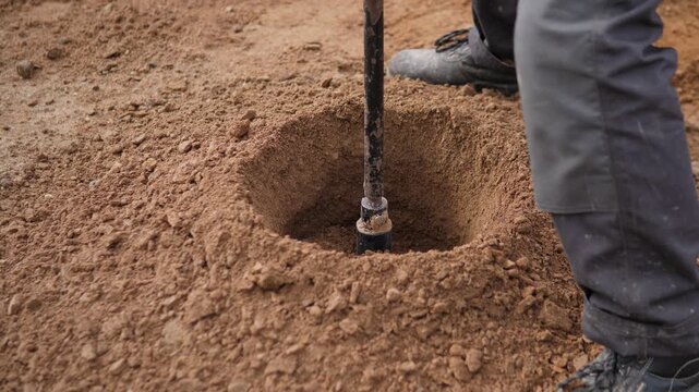 Ground-level view of worker using vertical digging tool in dry soil beside deep hole, with rough earth mound, compact dirt texture, and muted brown tones creating practical outdoor labor mood in
