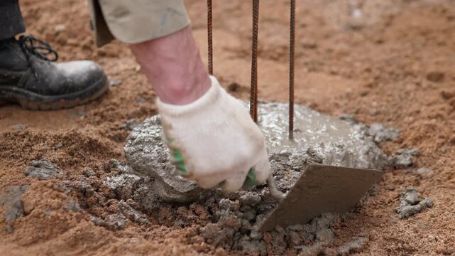 Close-up construction scene shows gloved hand shaping fresh wet concrete around vertical steel rebar with masonry trowel, with construction, concrete, rebar, trowel detail, showing glove, worker