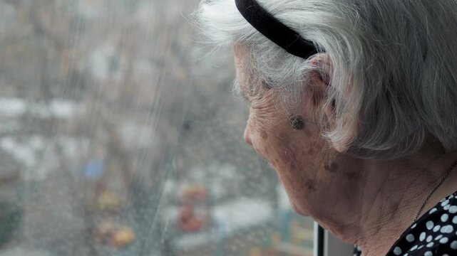 Pensive old woman with grey hair and deep wrinkles sadly looking out the window, feeling lonely and melancholic during a rainy day, expressing solitude and longing for memories