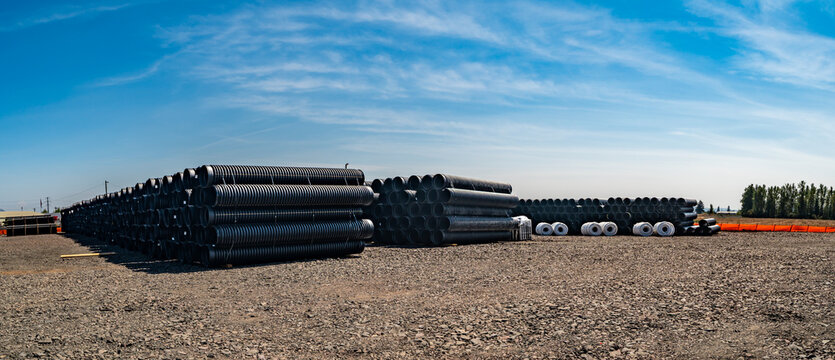 View of large stacks of black corrugated plastic drainage pipes stored in a gravel yard under a bright blue sky in Eugene, Oregon, United States.