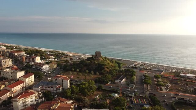 Aerial drone flight towards the Mediterranean sea over the coastal town of Scalea, Calabria, Italy, featuring the historic Torre Talao and beautiful beach at sunset, 4K.