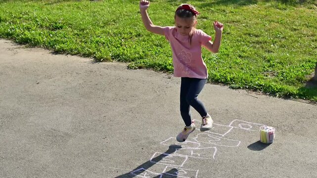 Preschool girl jumping along hopscotch grid drawn in sidewalk chalk on pavement under bright daylight. Great for outdoor recreation, childhood energy, and family clips. 4K slow motion footage