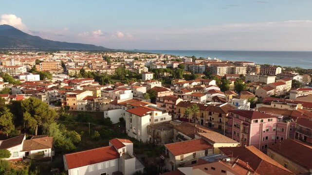 Aerial panning shot of the coastal town Scalea at golden hour, featuring traditional terracotta roofs, warm sunlight, and a dramatic mountain landscape in the background, Calabria, Italy, 4K.