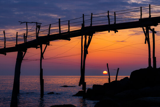 View of the traditional wooden trabocco fishing structure on the Costa dei Trabocchi at sunset with the sun dipping below the horizon over the Adriatic Sea in Marina di San Vito, Abruzzo, Italy.