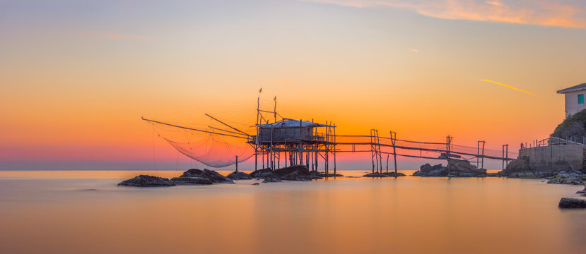 View of the Costa dei Trabocchi featuring a traditional wooden fishing structure over calm sea waters during a vibrant golden hour sunset in Marina di San Vito, Abruzzo, Italy.