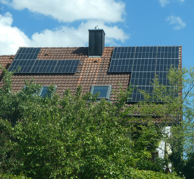 View of solar panels installed on a tiled roof with a chimney and skylights, set against a blue sky with clouds in Garching, Bavaria, Germany.