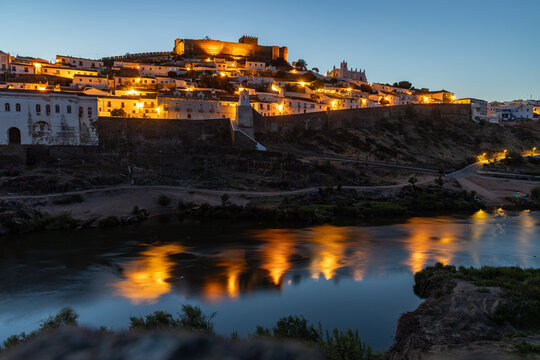 View of the illuminated historic town of Mertola and its hilltop castle reflecting in the calm waters of the Guadiana River in Mertola, Beja District, Portugal.