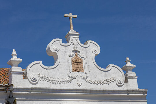 View of the ornate white pediment of a historic church featuring a stone cross and a carved coat of arms against a clear blue sky in Monsaraz, Evora District, Portugal.
