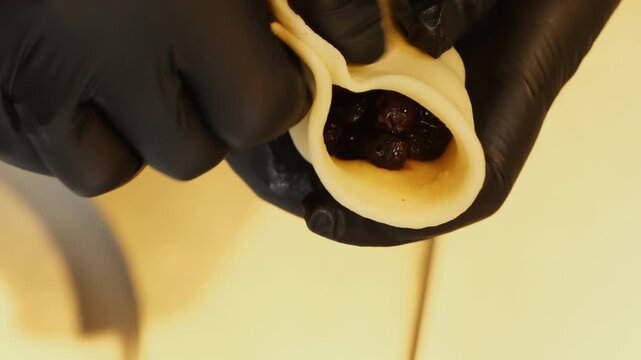 Gloved baker folds soft dough around dark berry filling inside pastry. Fingers bring edges together beginning sealing stuffed wrapper by hand
