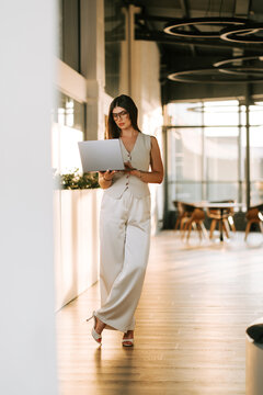 Professional woman in elegant office attire using a laptop while standing in a bright modern lobby