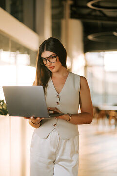 Young woman wearing glasses works on a silver laptop while standing inside a modern office space