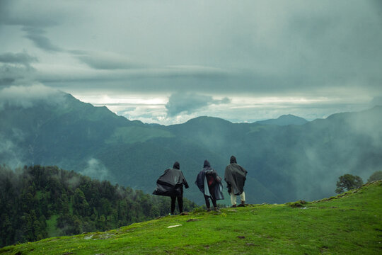 View of three hikers in rain ponchos standing on a grassy ridge overlooking the misty mountain ranges of the Ali Bedni Bugyal trek in Chamoli, Uttarakhand, India.