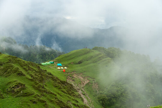View of colorful tents pitched on the lush green meadows of Ali Bedni Bugyal trek surrounded by misty clouds and rolling mountain slopes in Chamoli, Uttarakhand, India.