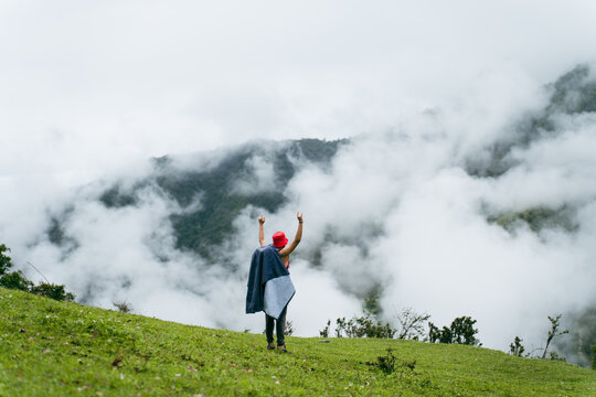 View of a hiker with a red hat and jacket standing on the grassy slopes of the Ali Bedni Bugyal trek with arms raised amidst thick mountain clouds in Chamoli, Uttarakhand, India.