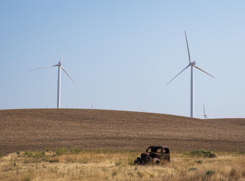 View of a rusted vintage car abandoned in a dry grassy field with large wind turbines standing on the rolling hills in the background in Oregon, United States.