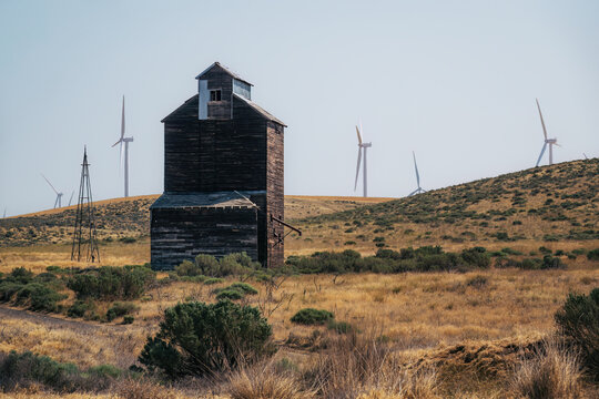 View of an old wooden grain elevator standing in a dry, grassy field in Eastern Oregon with wind turbines on the rolling hills in the background, Oregon, United States.
