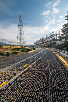 View of the metal grate roadway leading towards the Bonneville Dam with electrical towers and power infrastructure under a clear blue sky in Cascade Locks, Oregon, United States.
