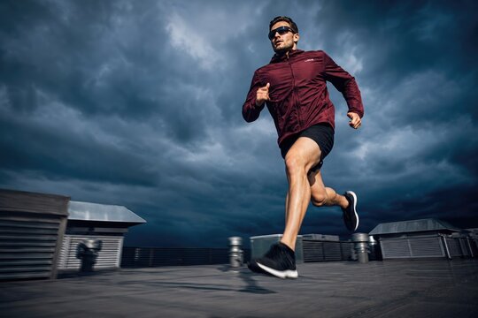 Determined male runner in maroon athletic jacket and black shorts sprinting on rooftop with dramatic stormy sky background, dynamic action shot, urban fitness concept