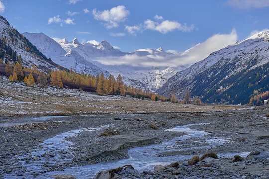 View of Engadin valley with snow-capped mountain peaks, autumn larch trees, and a winding river flowing through a rocky riverbed in Grisons, Switzerland.