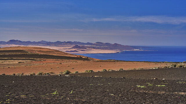 View of the volcanic landscape with dark soil, reddish-brown hills, and the blue ocean in the background in Lanzarote, Canarias, Spain.