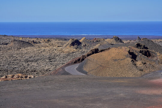 View of the volcanic landscape of Timanfaya National Park with dark lava fields, volcanic cones, and a winding road leading towards the blue Atlantic Ocean in Lanzarote, Canarias, Spain.
