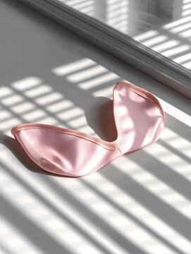 Pink silk sleep mask lies on a white surface. Sunlight casts shadows through a window. Minimalist, elegant, and luxurious feel