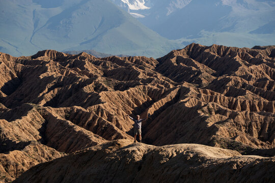 View of a person standing with arms raised on a ridge in the rugged, eroded landscape of the Ak-Say Valley with distant mountains, At-Bashi District, Naryn Region, Kyrgyzstan.