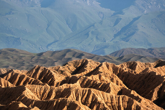 View of the rugged, golden rock formations of the Ak-Say Valley set against rolling green hills and distant blue mountains in At-Bashi District, Naryn Region, Kyrgyzstan.
