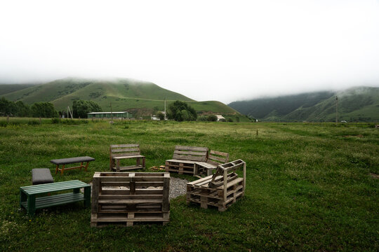 View of a rustic outdoor seating area made from wooden pallets arranged in a circle on a grassy field, with rolling green hills and low-hanging clouds in Tash-Bashat, Naryn Region, Kyrgyzstan.