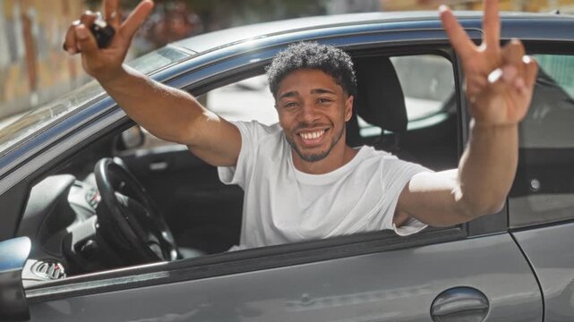 Young african american man in white t shirt leans from car window, holds keys and flashes thumbs up on street; joy success confidence.