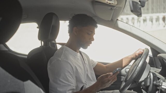 Man holding smartphone in right hand and steering with left while seated in a car on street, wearing white t shirt and seatbelt, looking at phone; concentration.