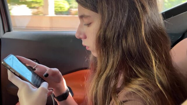 Focused teenager girl with long hair using her mobile phone for messaging and social media communication while traveling in a car through the city on a sunny day
