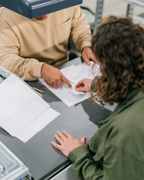 Hands of two people comparing blank documents on a dark charcoal desk with a task light for election integrity and verification processes