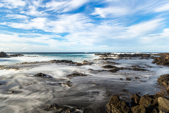 View of the rocky coastline with crashing waves and long exposure water motion under a bright blue sky with wispy clouds on Easter Island, Valparaiso, Chile.